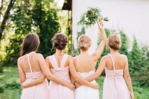 bride standing with her back in a white wedding dress and raised hand with a bouquet of protein and pistachio in the embraces of three bridesmaids in pink dresses transformer standing with her back and her hands down 