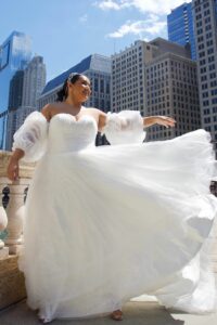 A beautiful bride wearing detachable sleeves for wedding dress with a billowing skirt and with tall buildings in the background.